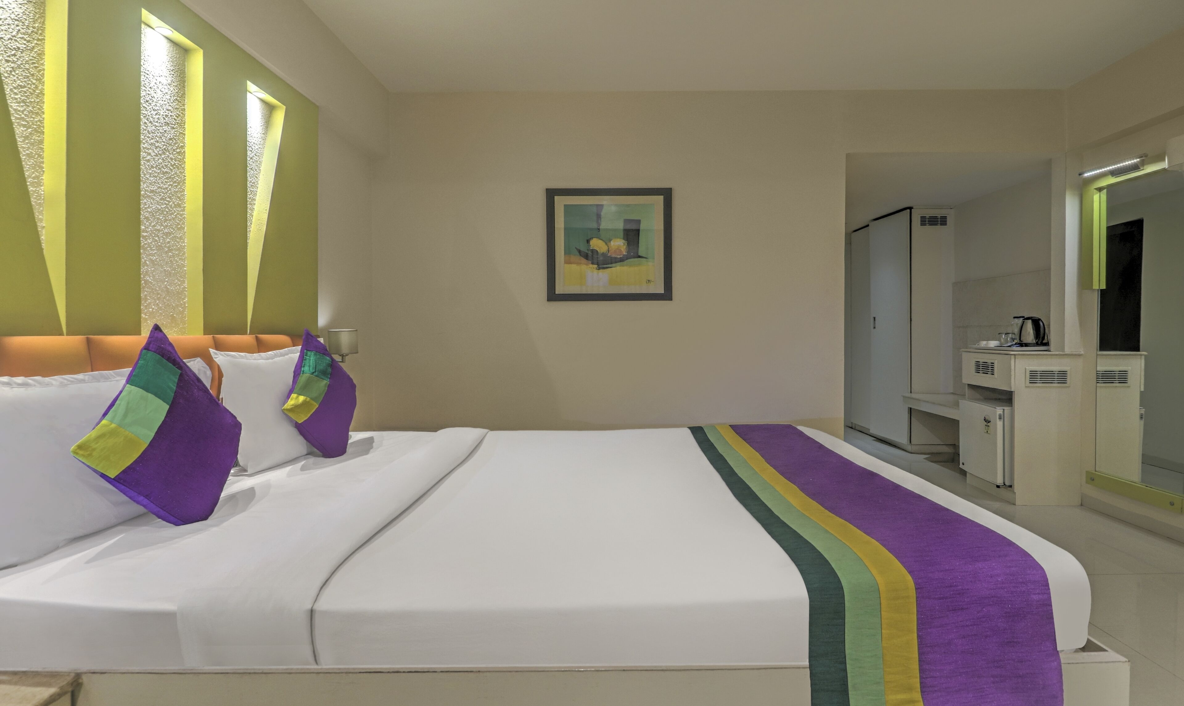 Ein Hotelzimmer mit Bett, einem Bild an der Wand, einem Schreibtisch mit Spiegel und einer Küchenzeile.
