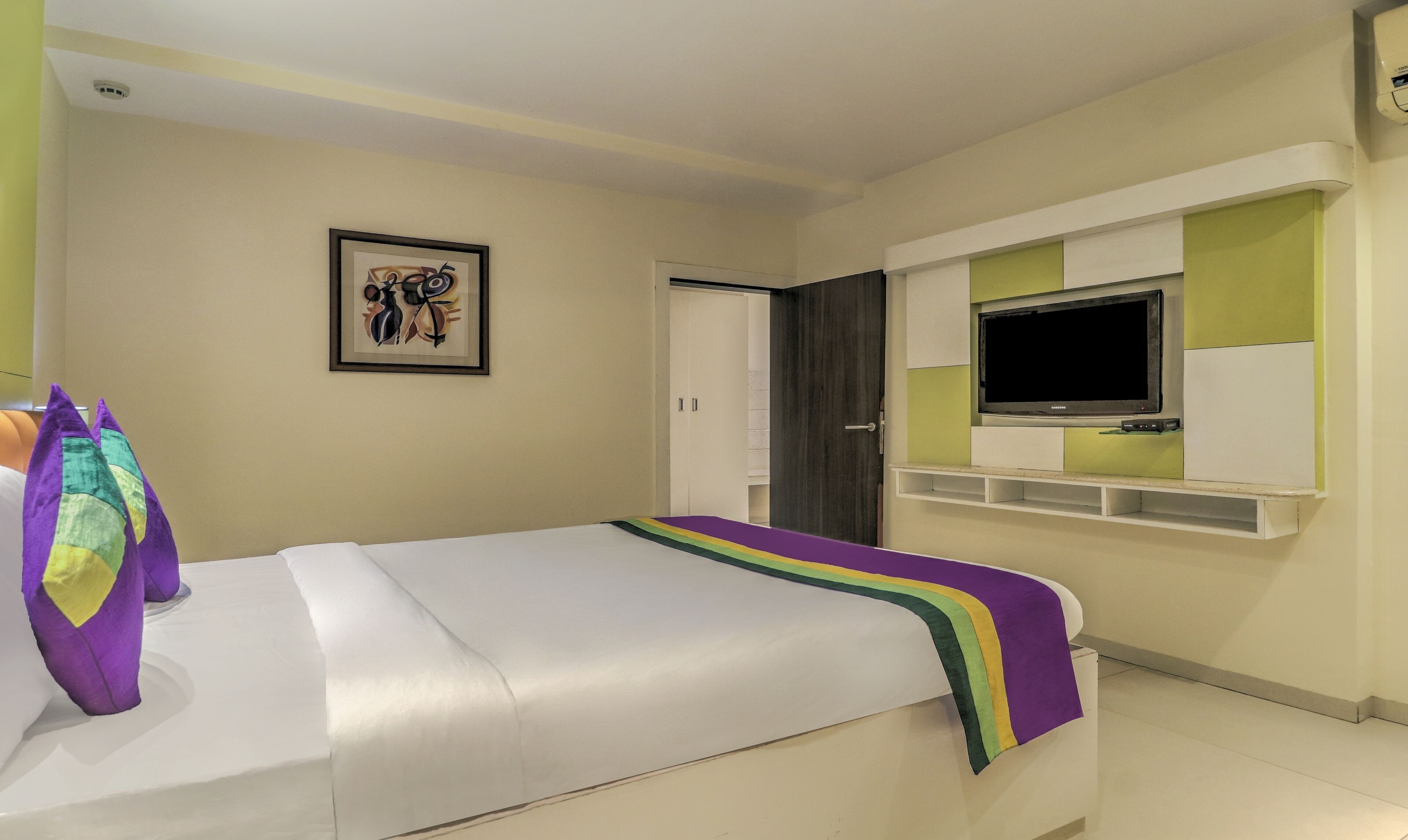 Ein Hotelzimmer mit Bett, Wandfernseher, Kleiderschrank und einem gerahmten Bild an der Wand.