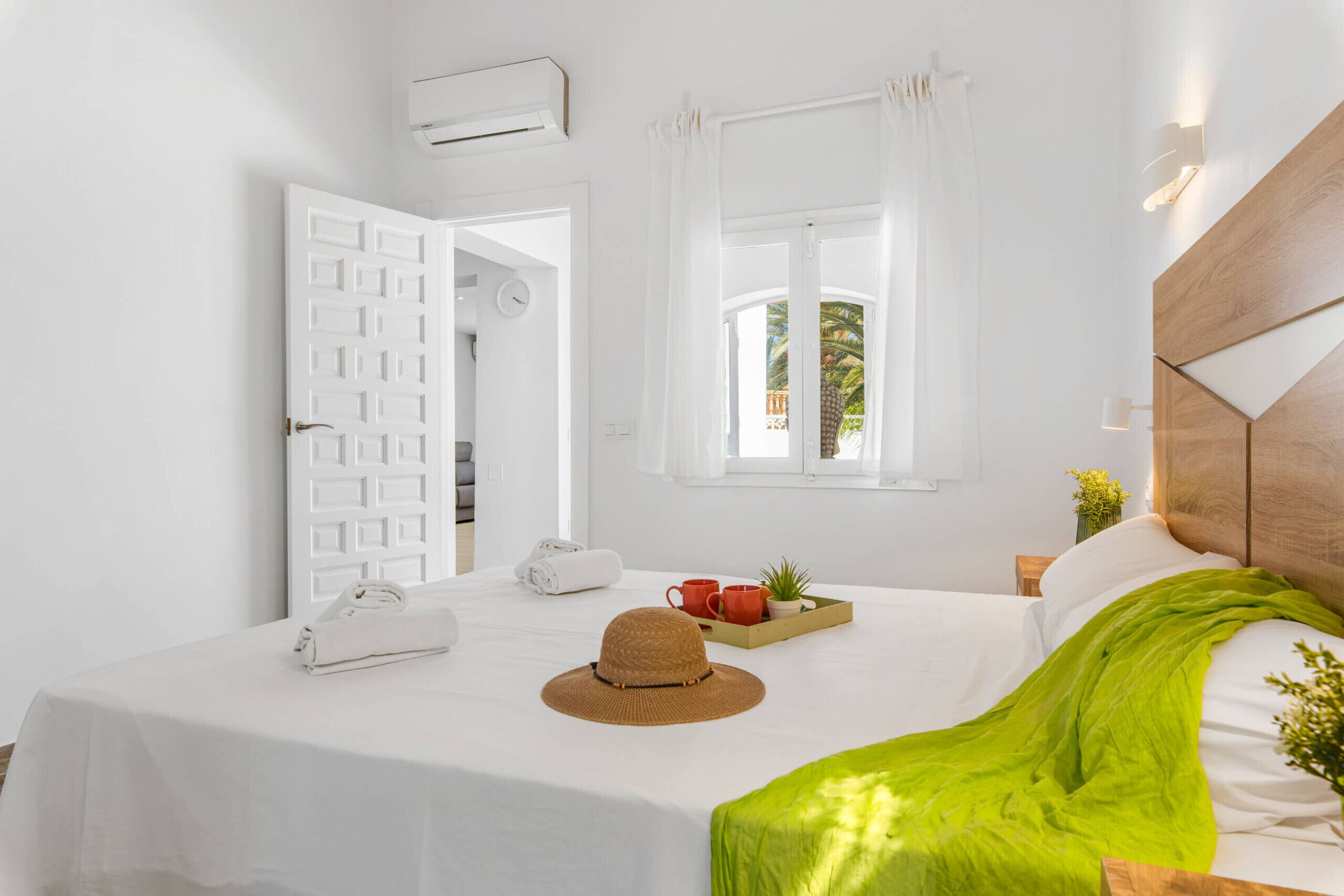 Habitación de hotel con cama, ventana con cortinas, aire acondicionado de pared y mesita de noche con un sombrero, una bandeja con tazas y una planta.