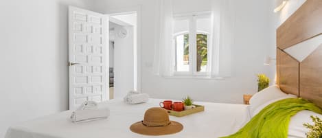 Habitación de hotel con cama, ventana con cortinas, aire acondicionado de pared y mesita de noche con un sombrero, una bandeja con tazas y una planta.