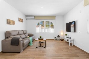Uma sala de estar moderna com um sofá, uma mesa de centro, uma televisão e uma janela com vista para palmeiras.
