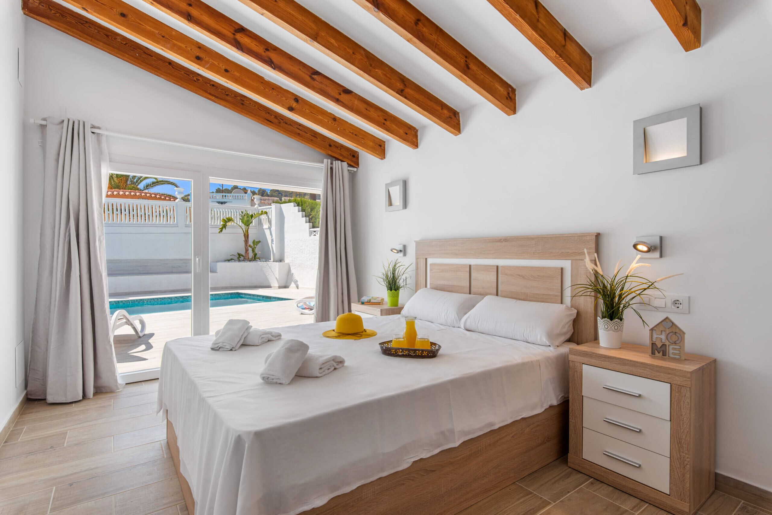 A hotel room with a bed, a bedside table, a nightstand, a window with curtains, and a view of a pool and outdoor area.