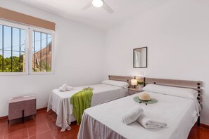 Habitación de hotel con dos camas, una mesita pequeña, un banco, un ventilador de techo y una ventana con vistas a la vegetación.