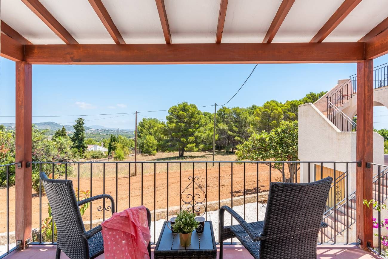 Un balcon avec des fauteuils en osier, une petite table ornée d’une plante et une vue sur un paysage rural.