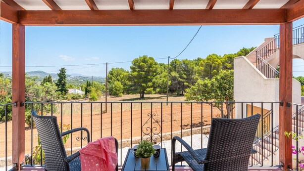 Uma varanda com cadeiras de vime, uma pequena mesa com uma planta e vista para uma paisagem rural.