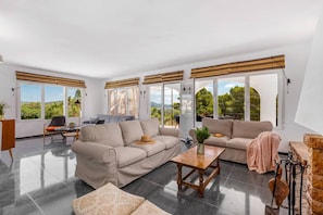 A spacious living area with a sofa set, a coffee table, and a view of greenery through large windows.