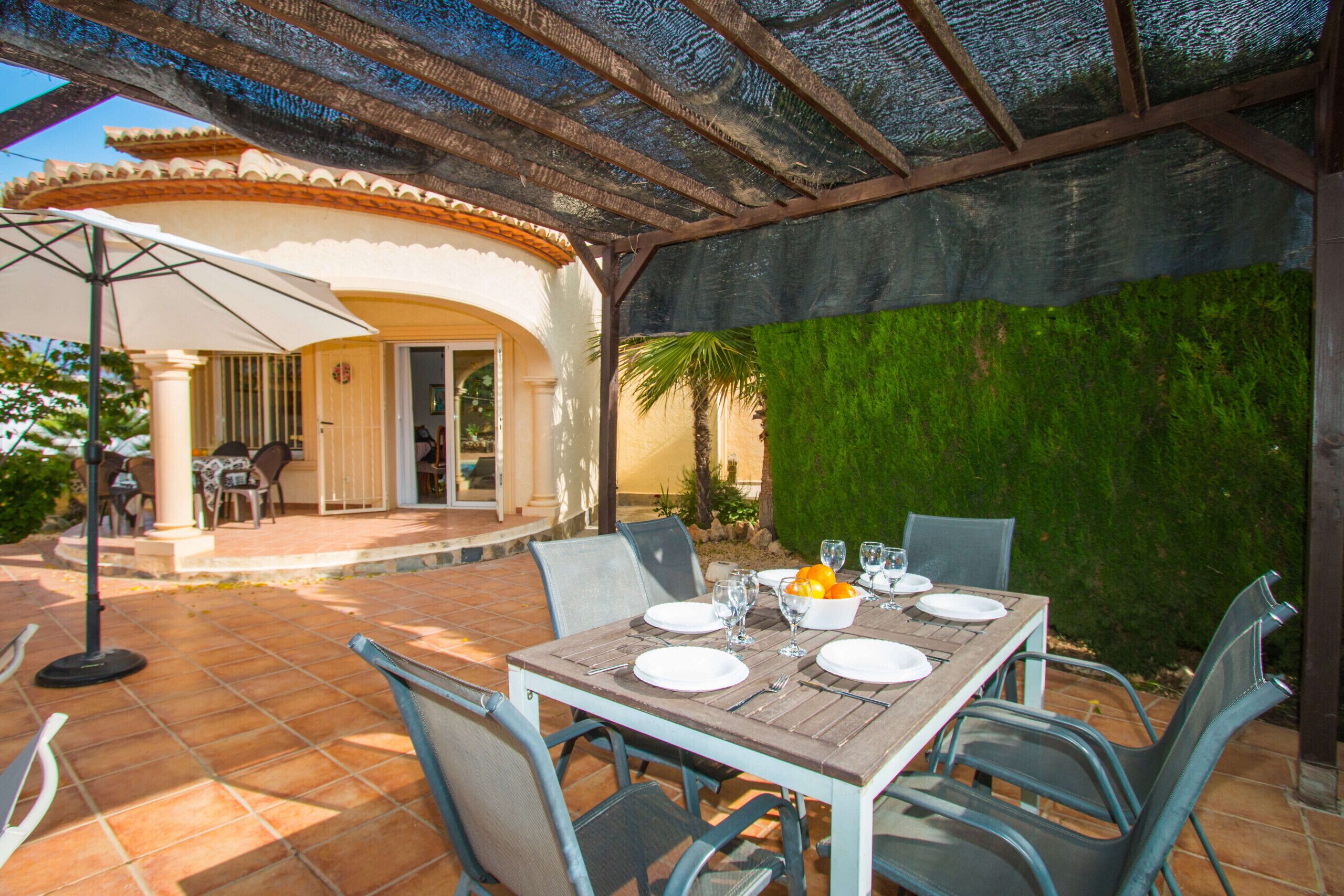 An outdoor patio with a table set for dining, surrounded by chairs, and a covered area with a pergola overhead.