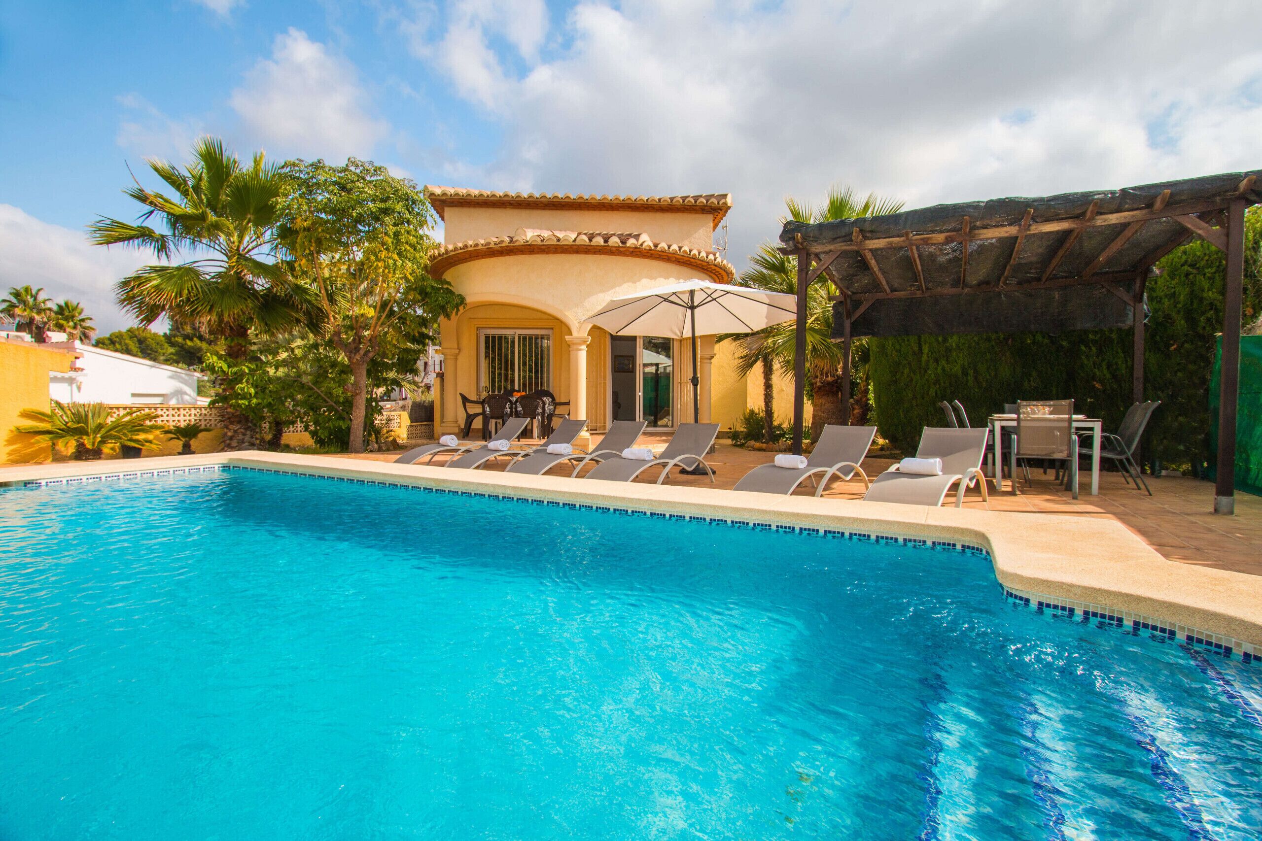 A swimming pool with lounge chairs and umbrellas, a covered patio area, and a building with a tiled roof.