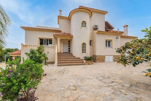 Uma casa de dois andares com um pátio pavimentado em pedra, uma escadaria central e um pequeno jardim com plantas floridas.