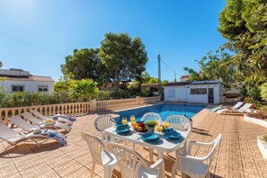 Área de piscina com espreguiçadeiras, uma mesa posta para refeição e um pequeno edifício ao fundo.