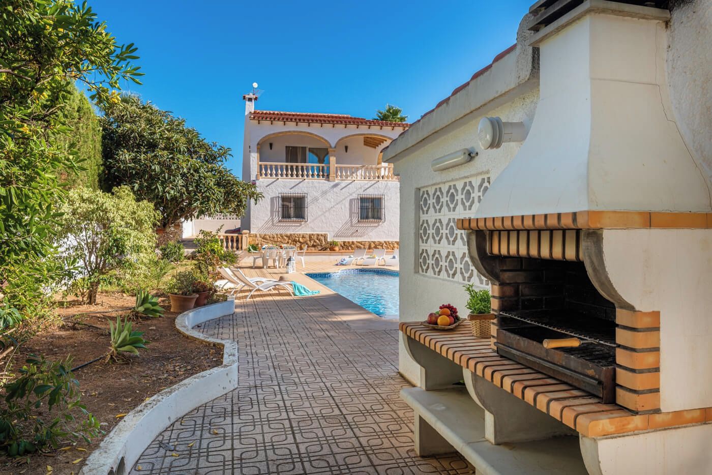 A pool area with a stone-tiled deck, a barbecue grill, and a view of a villa with a balcony.
