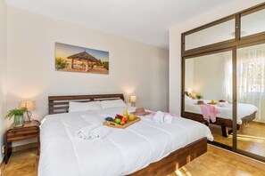 Quarto de hotel com uma cama grande, mesa de cabeceira com abajur, bandeja de frutas e um quadro na parede.