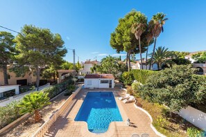 Piscina com deck de azulejos, cercada por vegetação e um pequeno edifício com telhado em tijolos vermelhos.