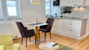Moderni keittiö, jossa on valkoiset kaapit, mikroaaltouuni ja erilaisia keittiövälineitä. Keittiössä on ruokailutila, jossa on pöytä ja tuolit, sekä pieni pyöreä pöytä, jonka päällä on papereita ja kaukosäädin.