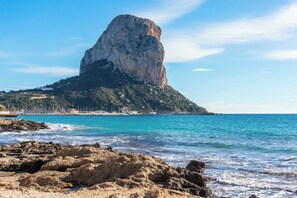 Paisagem costeira com uma formação rochosa imponente, mar azul cristalino e uma praia arenosa.