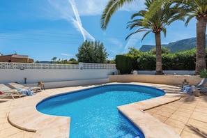 A swimming pool with a curved edge, surrounded by lounge chairs and palm trees. - Villa Reva - PlusHolidays (Calpe)