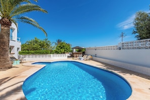 Piscina com parede branca e palmeiras, cercada por um muro branco e um prédio com fachada branca.
