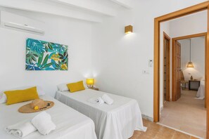 A hotel room with two single beds, a wall-mounted air conditioner, a painting, a bedside lamp, and a door leading to another room.