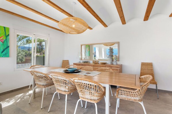 A dining area with a wooden table and wicker chairs, a hanging light fixture, and a large mirror above the table.