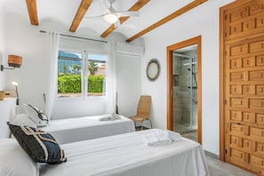Quarto de hotel com duas camas, chuveiro, vista para o verde e porta de madeira.
