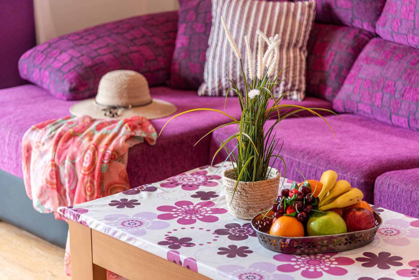 A cozy living room with a purple sofa, a woven plant pot, and a fruit bowl on a table with a floral tablecloth.