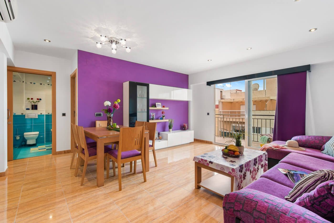 Ein modernes Wohnzimmer mit einer lilafarbenen Akzentwand, einem Essbereich mit Holzmöbeln und einem Balkon mit Blick.