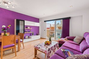 A modern living space with a purple accent wall, a dining area with wooden furniture, a sofa set, and a balcony with a view.