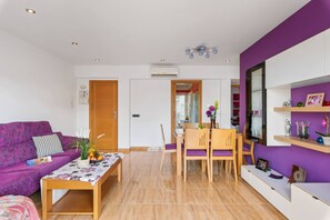 Sala de estar moderna com sofá roxo, mesa de jantar de madeira e área de cozinha visível através de uma porta aberta.