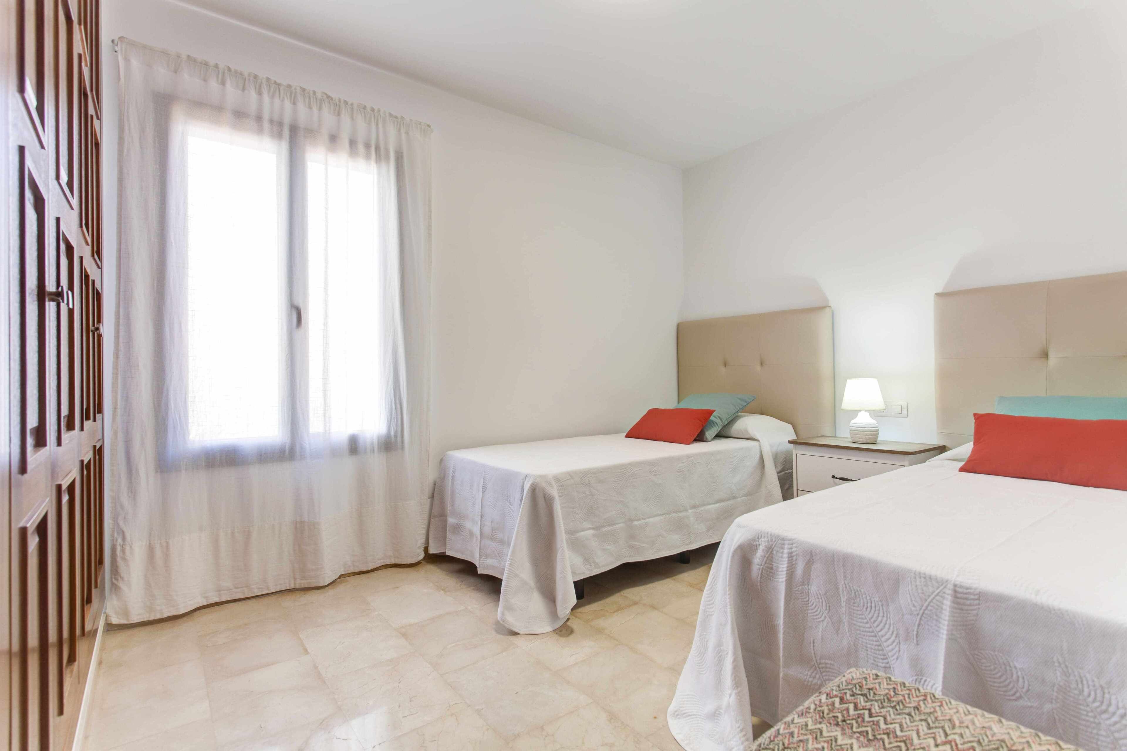 A hotel room with two beds, a window with curtains, a bedside table with a lamp, and a tiled floor.