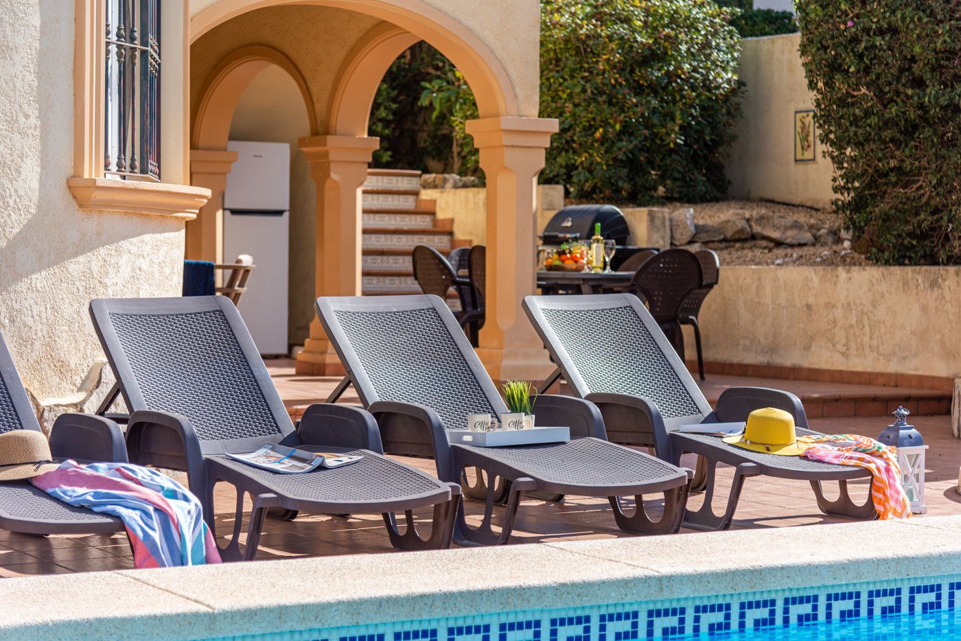 Un espace aménagé au bord de la piscine, avec des chaises longues, une table sur laquelle se trouvent un chapeau et une lanterne, et un barbecue à l’arrière-plan.
