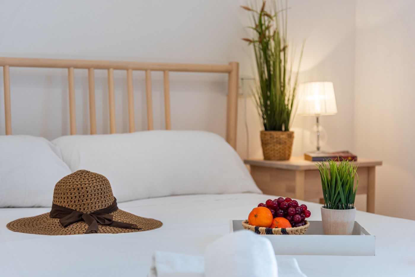 Una cama bien tendida con sábanas blancas, un sombrero tejido sobre la cama, una mesita de noche con una lámpara, una planta en maceta y una bandeja de frutas.