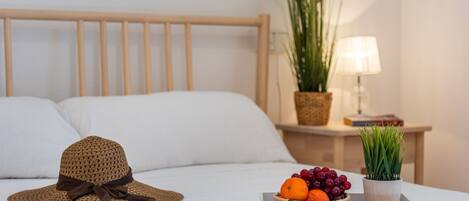 Una cama bien tendida con sábanas blancas, un sombrero tejido sobre la cama, una mesita de noche con una lámpara, una planta en maceta y una bandeja de frutas.