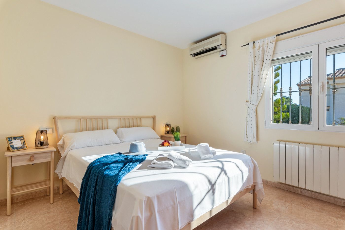 Une chambre à coucher comprenant un lit, des tables de chevet, une commode, un appareil de climatisation, une fenêtre avec des rideaux et une vue sur la verdure à l’extérieur.
