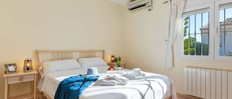 Un dormitorio con cama, mesitas de noche, unidad de aire acondicionado, ventana con cortinas y vistas a zonas verdes.