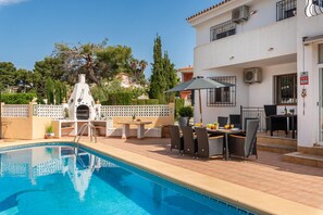 A swimming pool with a sun lounger and outdoor furniture, a white building with balconies, and a clear sky.