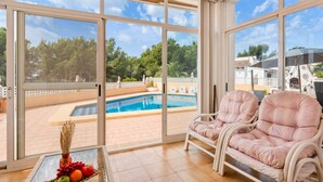 Una habitación iluminada por el sol con vistas a una piscina, dos sillones cómodos y una mesa con fruta fresca.