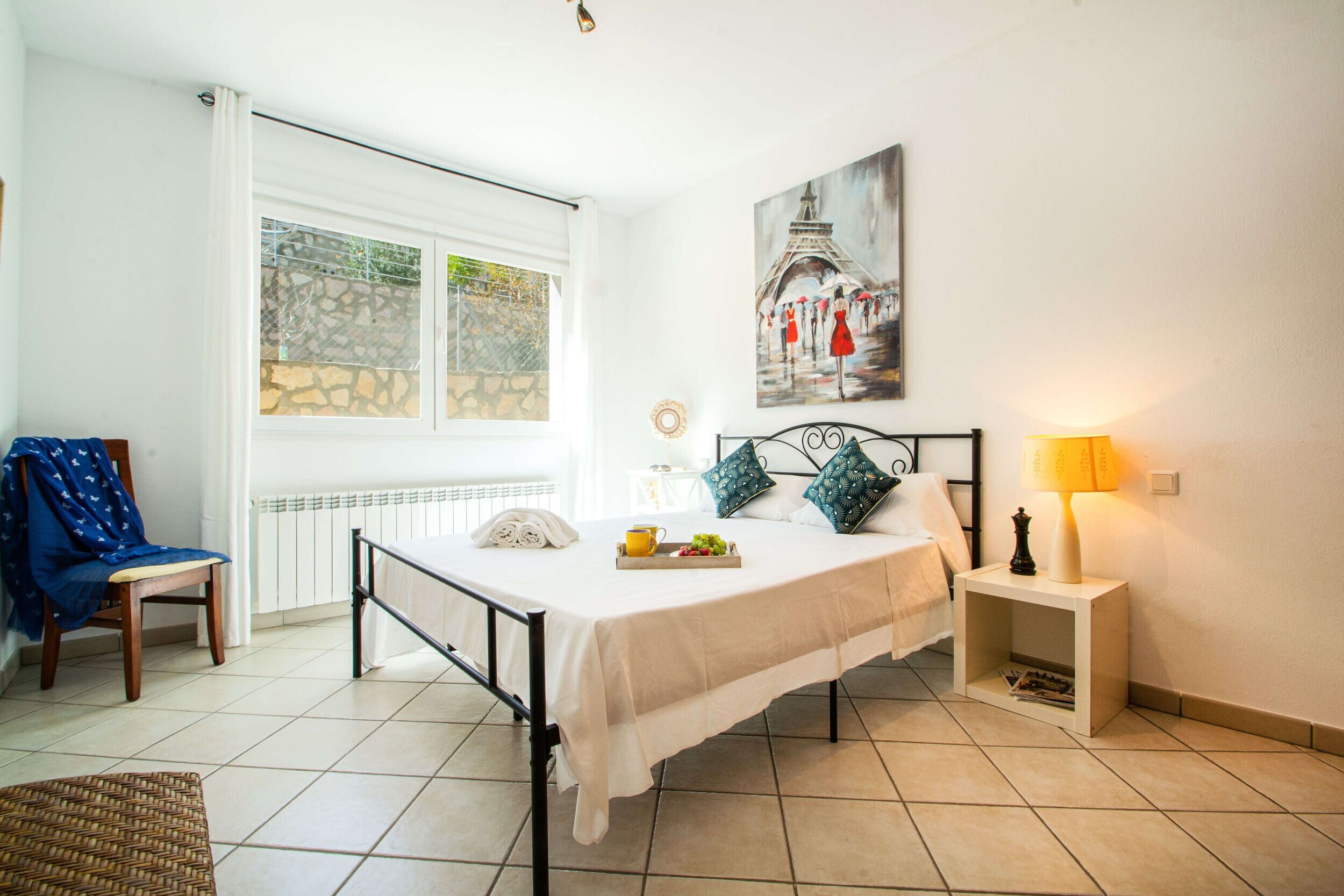 A bedroom with a bed, a chair, a bedside table, a lamp, a painting, and a window with a view of a stone wall.