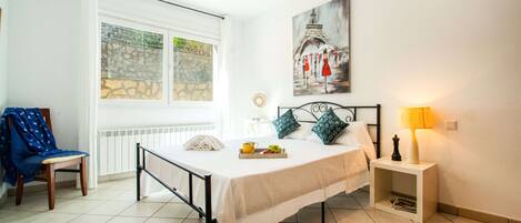 A bedroom with a bed, a chair, a bedside table, a lamp, a painting, and a window with a view of a stone wall.