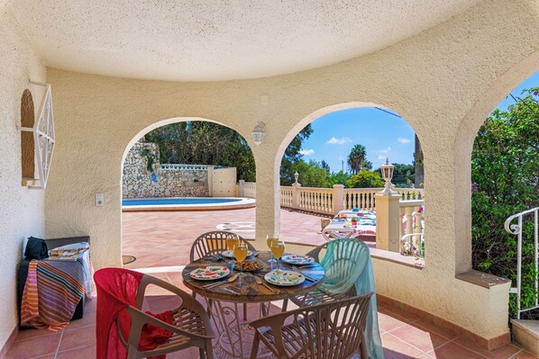 A covered outdoor dining area with a pool view, featuring a table set for two with plates, glasses, and cutlery.