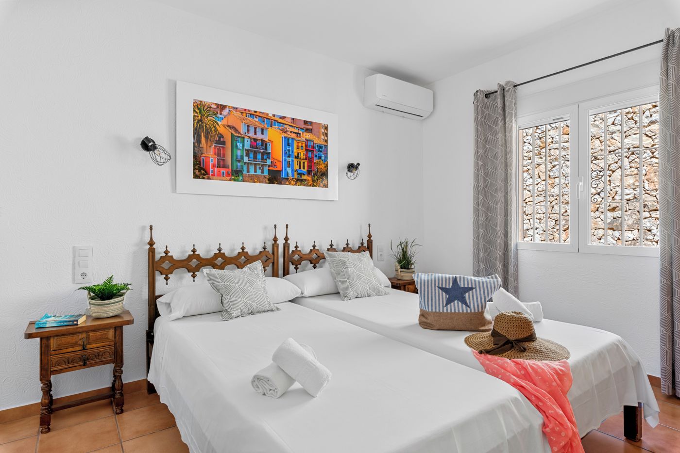 A hotel room with two beds, a painting, an air conditioning unit, a window with curtains, and a small table with a plant.