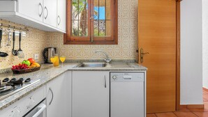 Cozinha compacta com armários brancos, lava-louças embutida, pia e janela com vista para palmeiras.