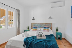 A hotel room with a bed, a window, a wall-mounted air conditioner, and a small table with a plant.