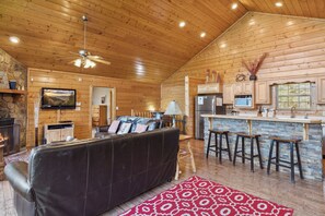 A cozy cabin interior with a fireplace, a leather sofa, a dining area with stools, and a kitchen with wooden cabinets.