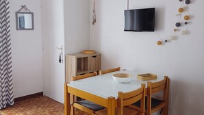 A dining area with a wooden table and chairs, a hanging light fixture, a television mounted on the wall, and a patterned rug on the floor.