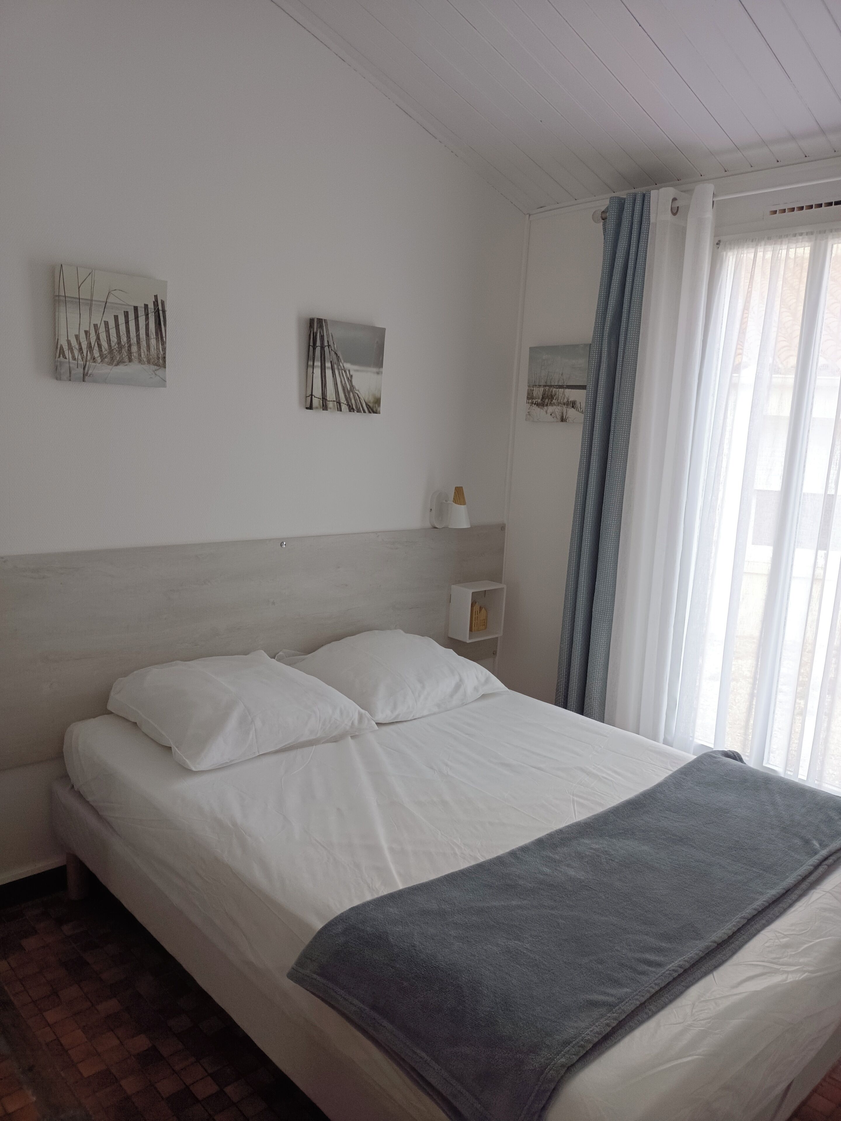 Una cama bien hecha con ropa blanca y una manta gris, una mesita de noche blanca con una lámpara y dos cuadros en la pared que muestran paisajes naturales.