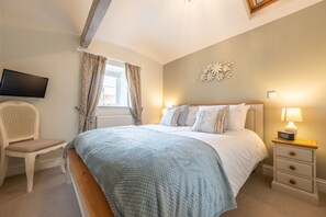 A bedroom with a bed, a nightstand with a lamp, a chair, a TV mounted on the wall, and a window with curtains.