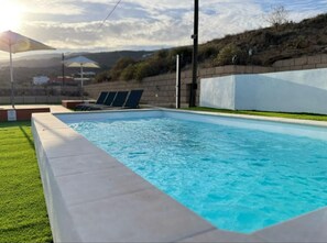 A swimming pool with a clear blue water surface, surrounded by a low concrete edge. There are lounge chairs along the poolside, and a grassy area in the foreground.