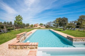 Una piscina rectangular con bordes de piedra y ladrillo, rodeada por un césped con árboles y sillas de descanso.