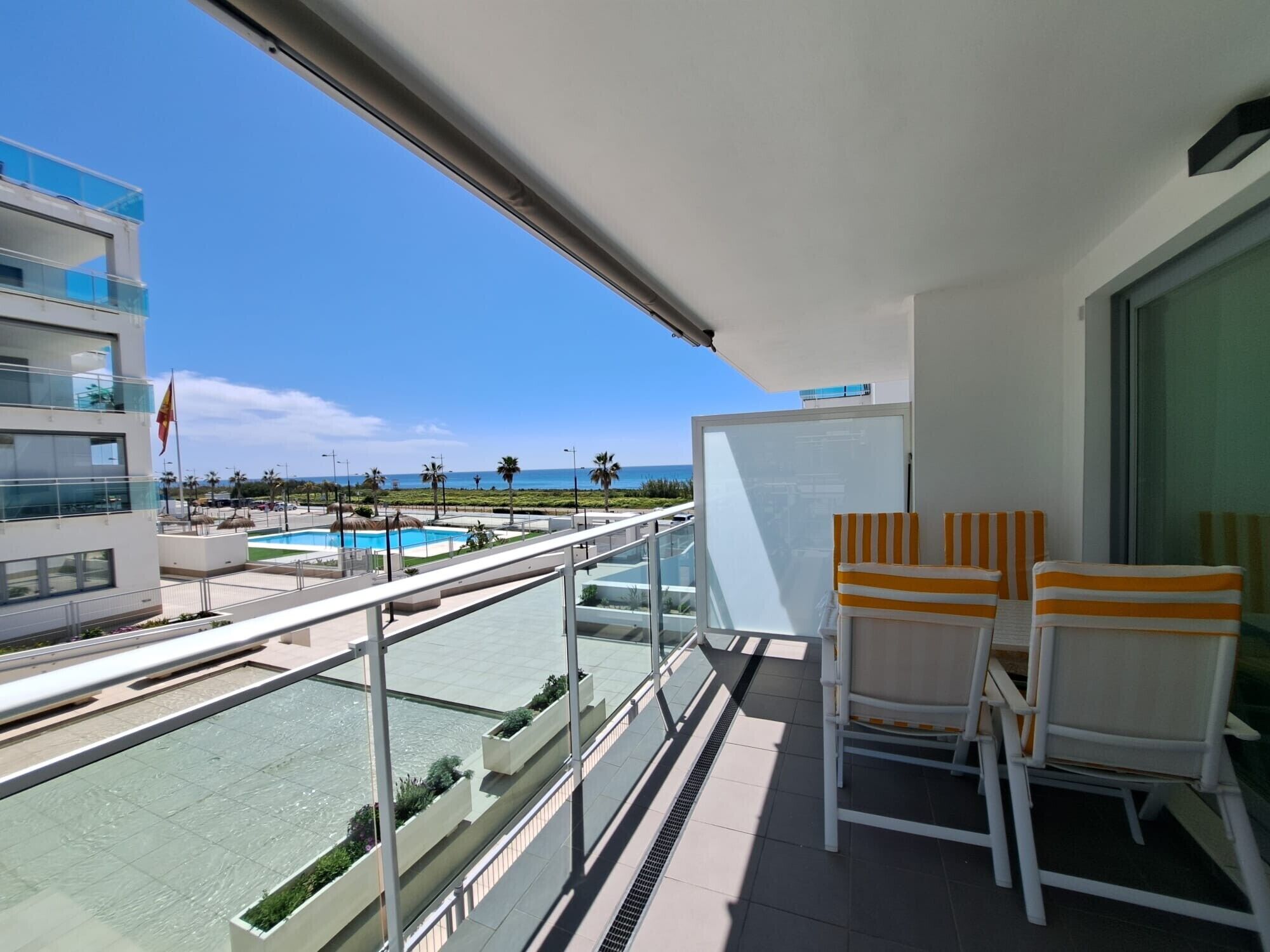 A balcony with a view of a pool and beach, two striped chairs, and a whiteboard.