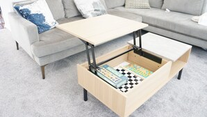 Un salon moderne avec un canapé gris, une table basse en bois surmontée d’un jeu d’échecs et une petite commode en bois.